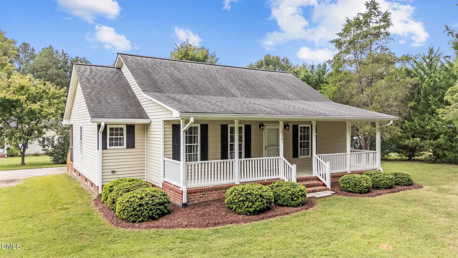 Charming single-story beige house with white trim at 6581 Hwy 158 Bus, Henderson. Features a black shingle roof, covered front porch with white railings, manicured shrubs, lush green grass, and a tree-lined yard.