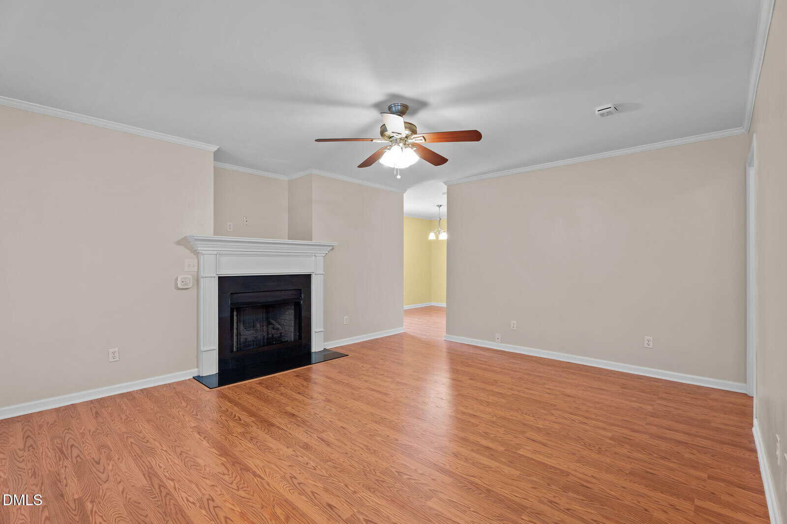A spacious living room with light beige walls, hardwood floors, a white fireplace with a black hearth, a white ceiling with a ceiling fan, and recessed lighting. An adjacent room is visible through an open doorway.