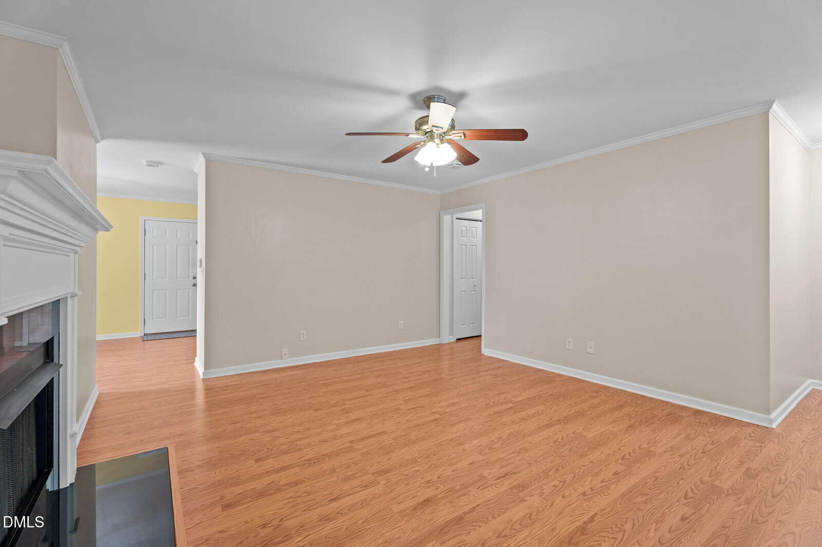 A spacious living room with light beige walls, white trim, a ceiling fan with lights, wood flooring, a white mantel fireplace on the left, and multiple doorways leading to other rooms.
