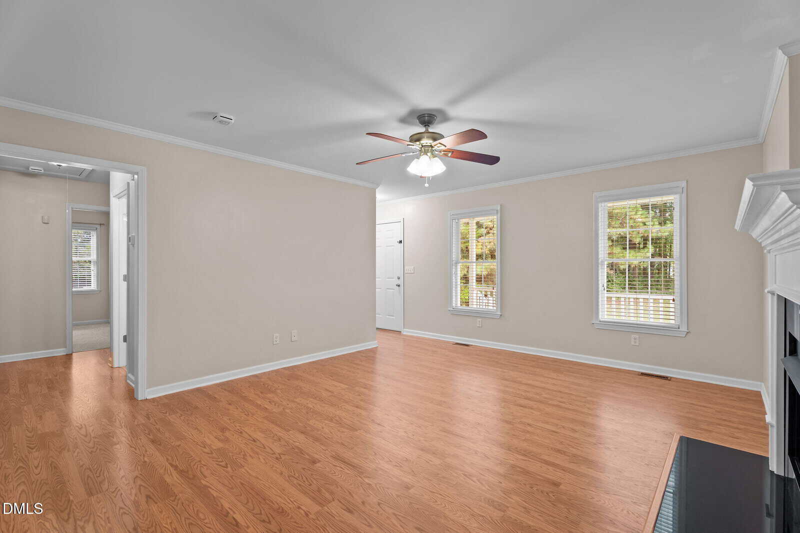 A spacious living room with light beige walls, hardwood floors, a ceiling fan with lights, two windows, and a white fireplace. There are doorways leading to other rooms and white trim along the ceiling and floors.
