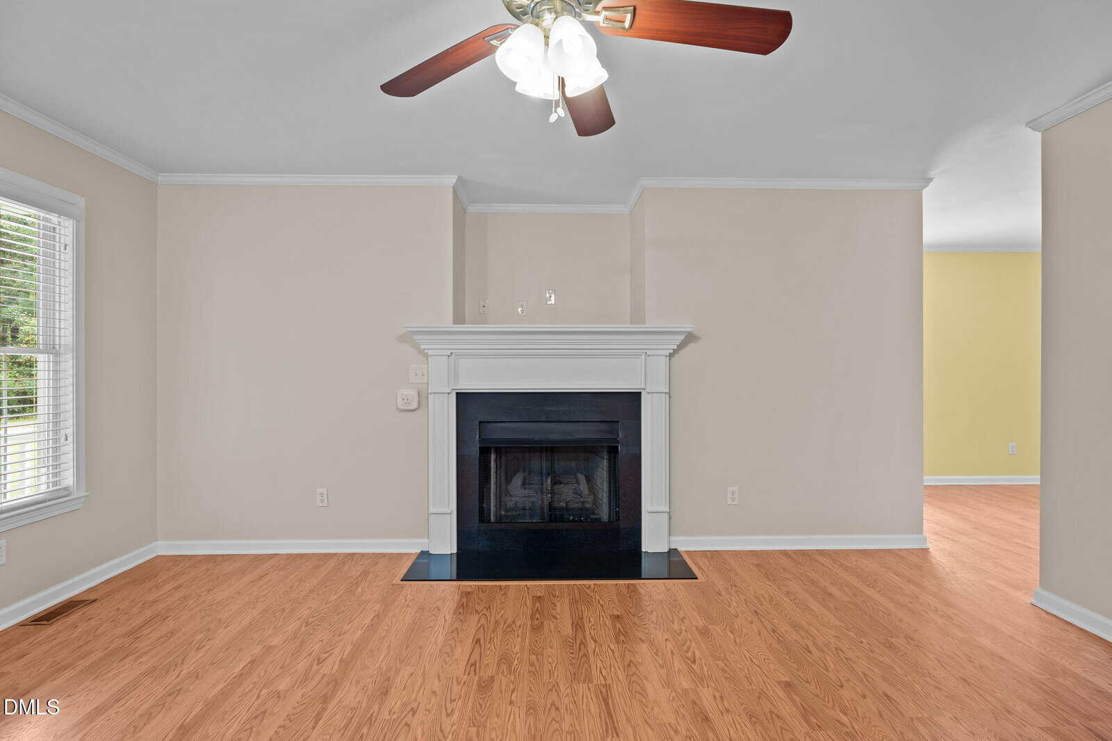 A living room with light beige walls, hardwood floors, a black fireplace with a white mantel, ceiling fan with lights, and a large window with white blinds letting in natural light.