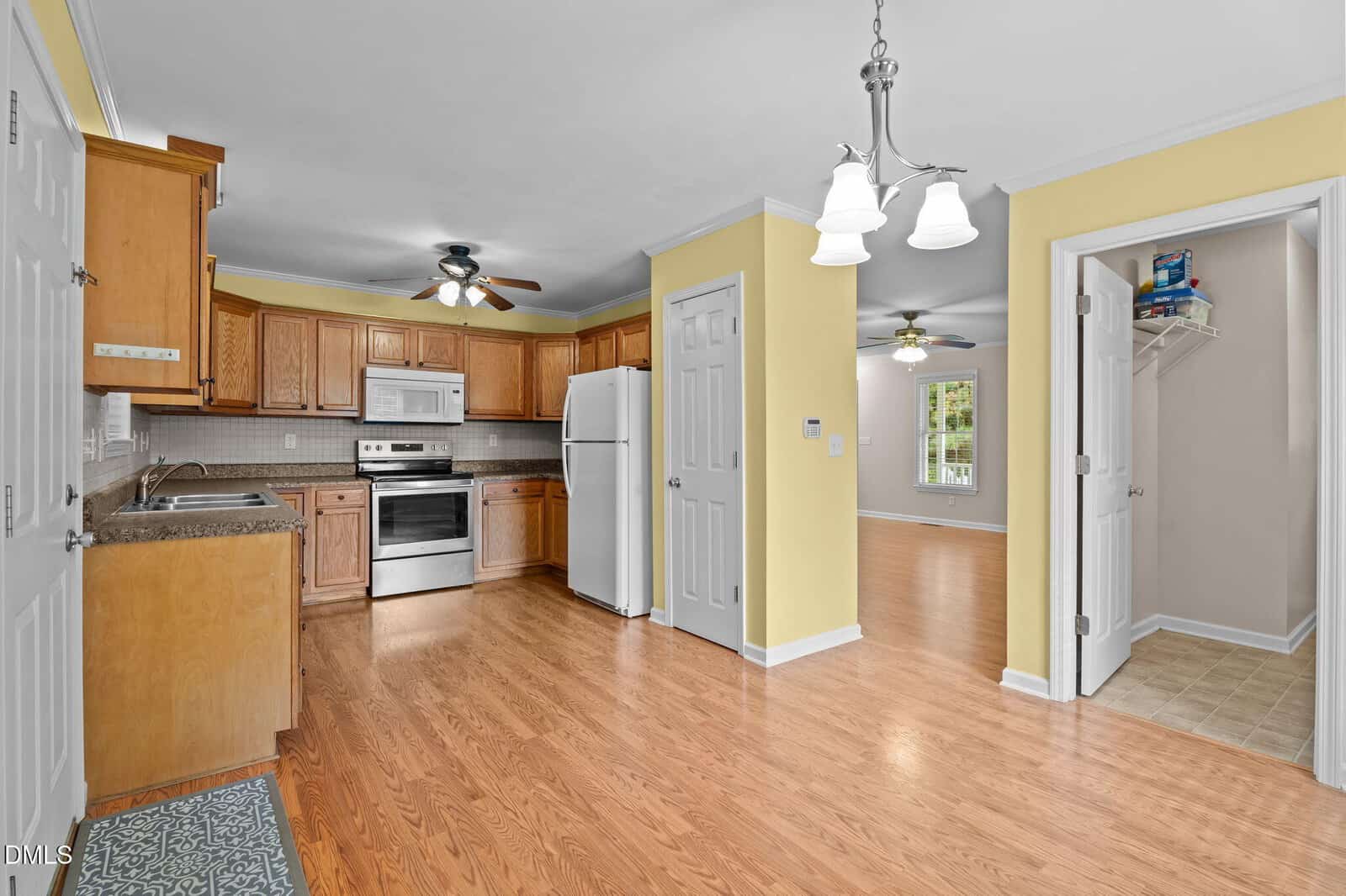 A kitchen with light wood cabinets, stainless steel appliances, white refrigerator, and double basin sink. The floor is light wood, walls are yellow, and adjacent rooms are visible through open doorways.
