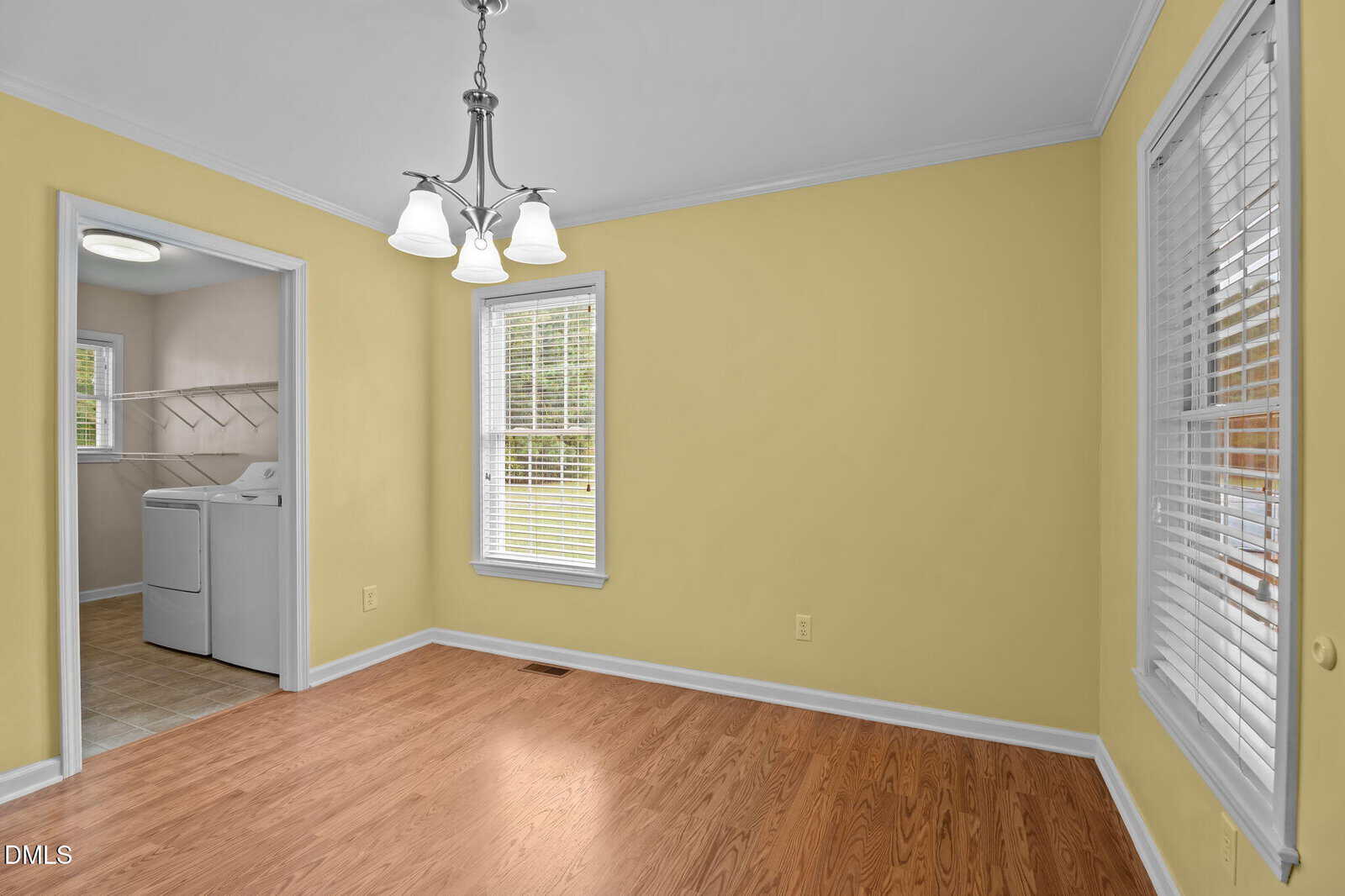 A bright room with yellow walls, wood flooring, two windows with blinds, and a ceiling light fixture. A laundry room with a washer and dryer is visible through an open doorway.