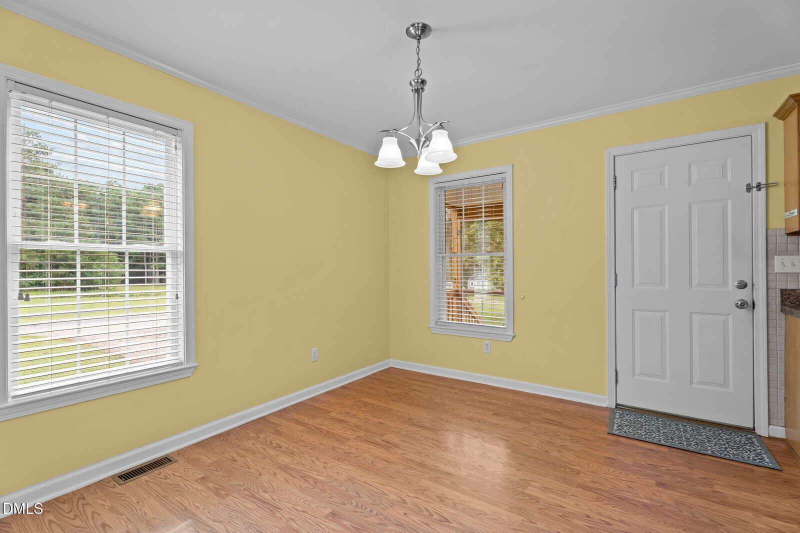 A small, empty room with yellow walls, light hardwood flooring, two windows with white blinds, a white door, and a ceiling light fixture with three lamps.