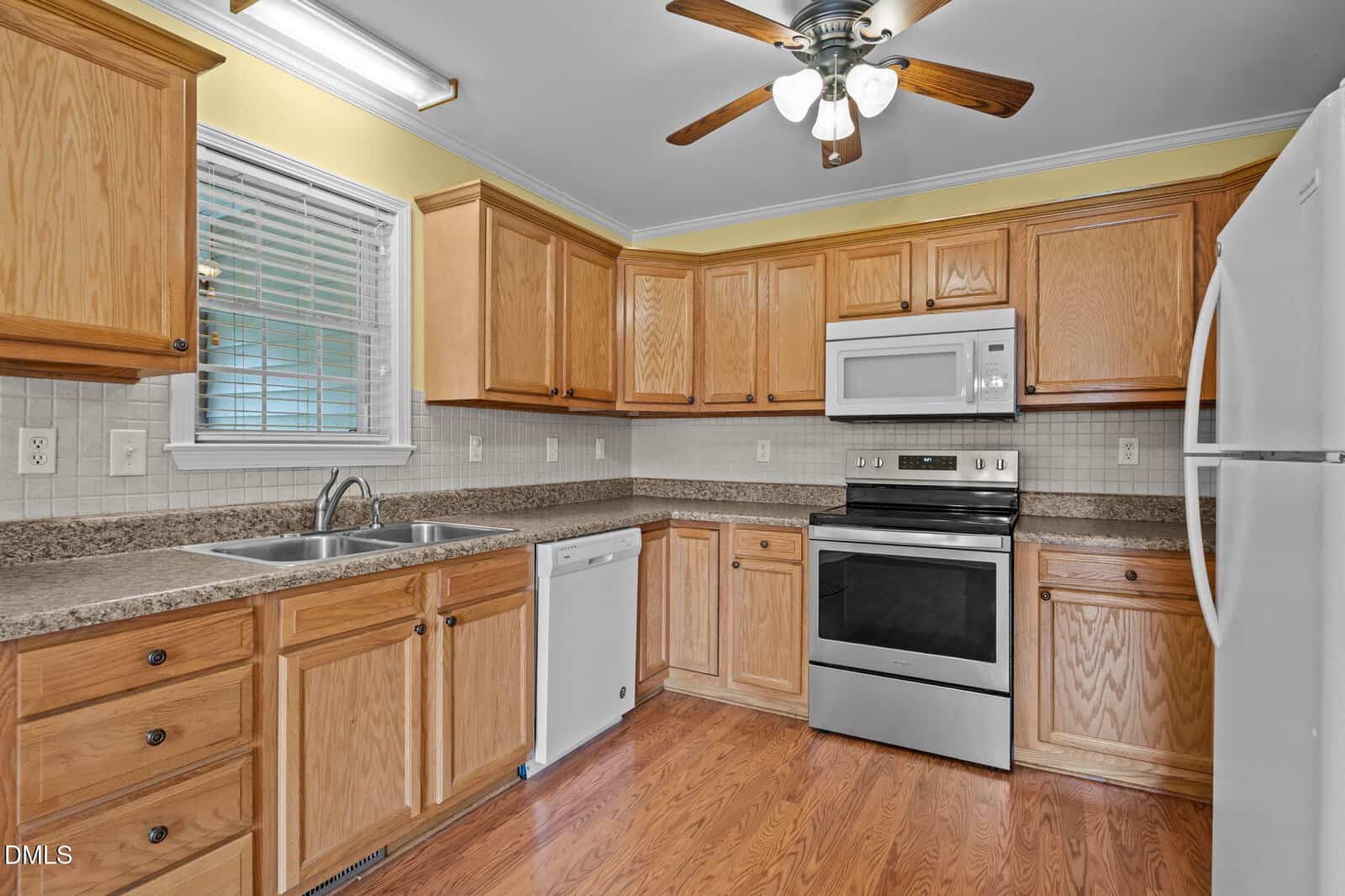 A kitchen with wood cabinets, granite countertops, stainless steel oven and microwave, white refrigerator and dishwasher, double sink under a window, tile backsplash, wood flooring, and a ceiling fan with lights.