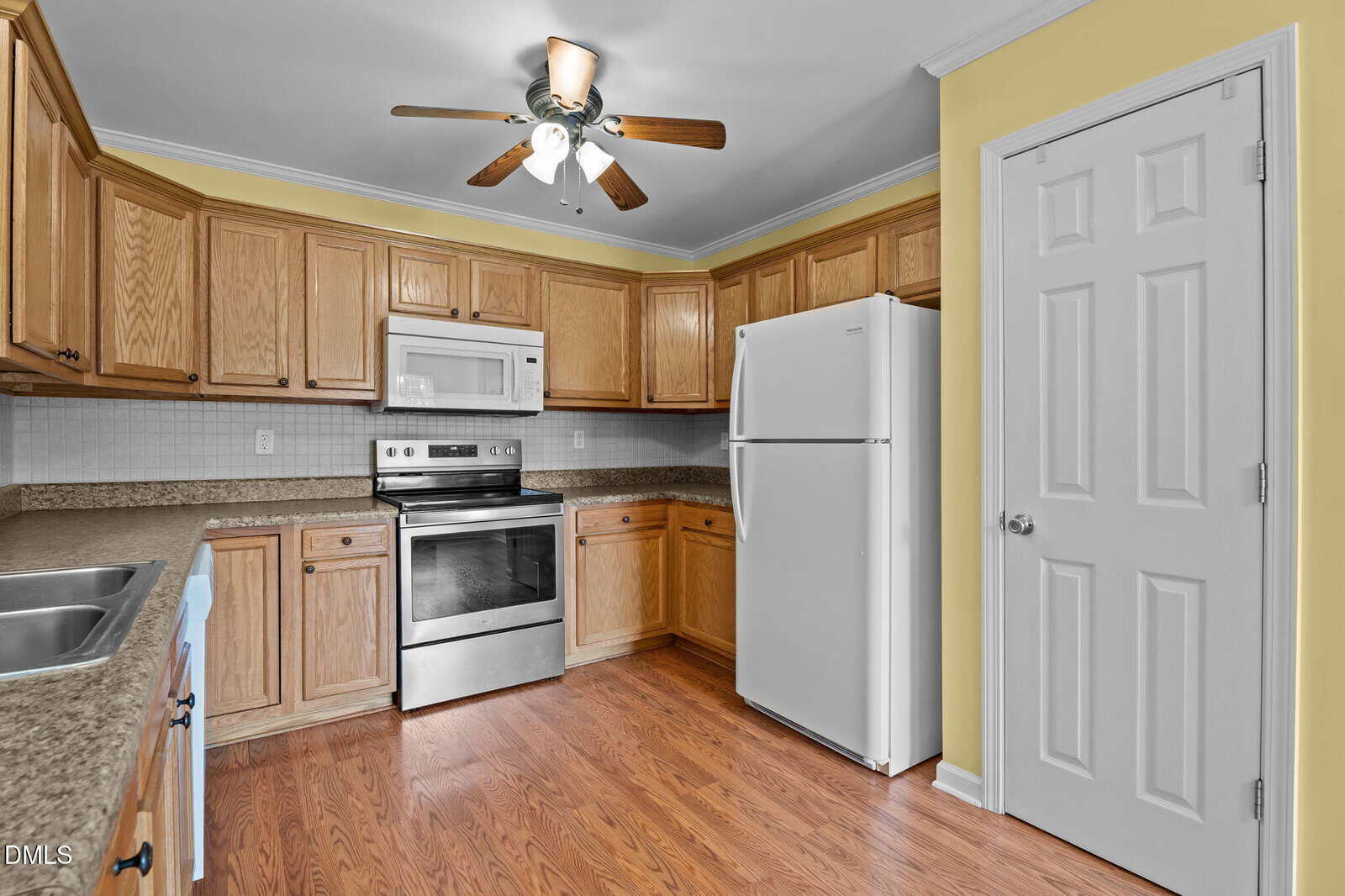 Kitchen with wooden cabinets, white refrigerator, microwave, electric stove, stainless steel sink, brown countertops, and wood-look flooring. Ceiling fan and light fixture overhead, walls painted yellow, door to the right.
