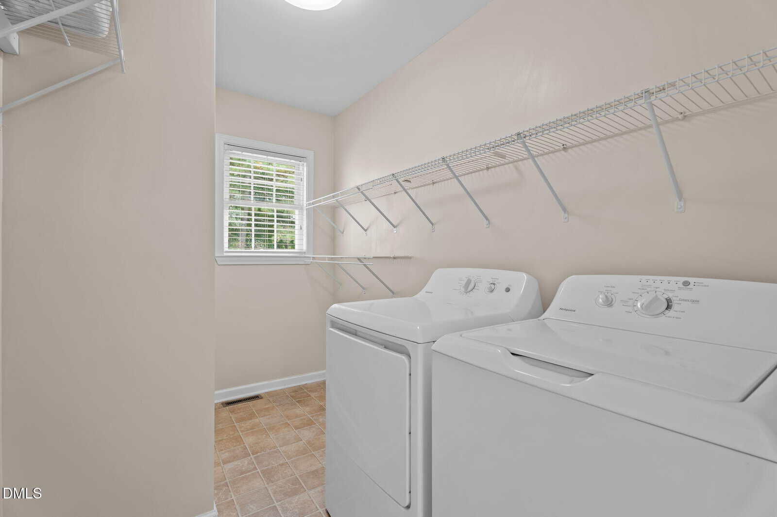 Laundry room with a washer and dryer, beige walls, tile floor, a window with white blinds, and two white wire shelves along the walls.