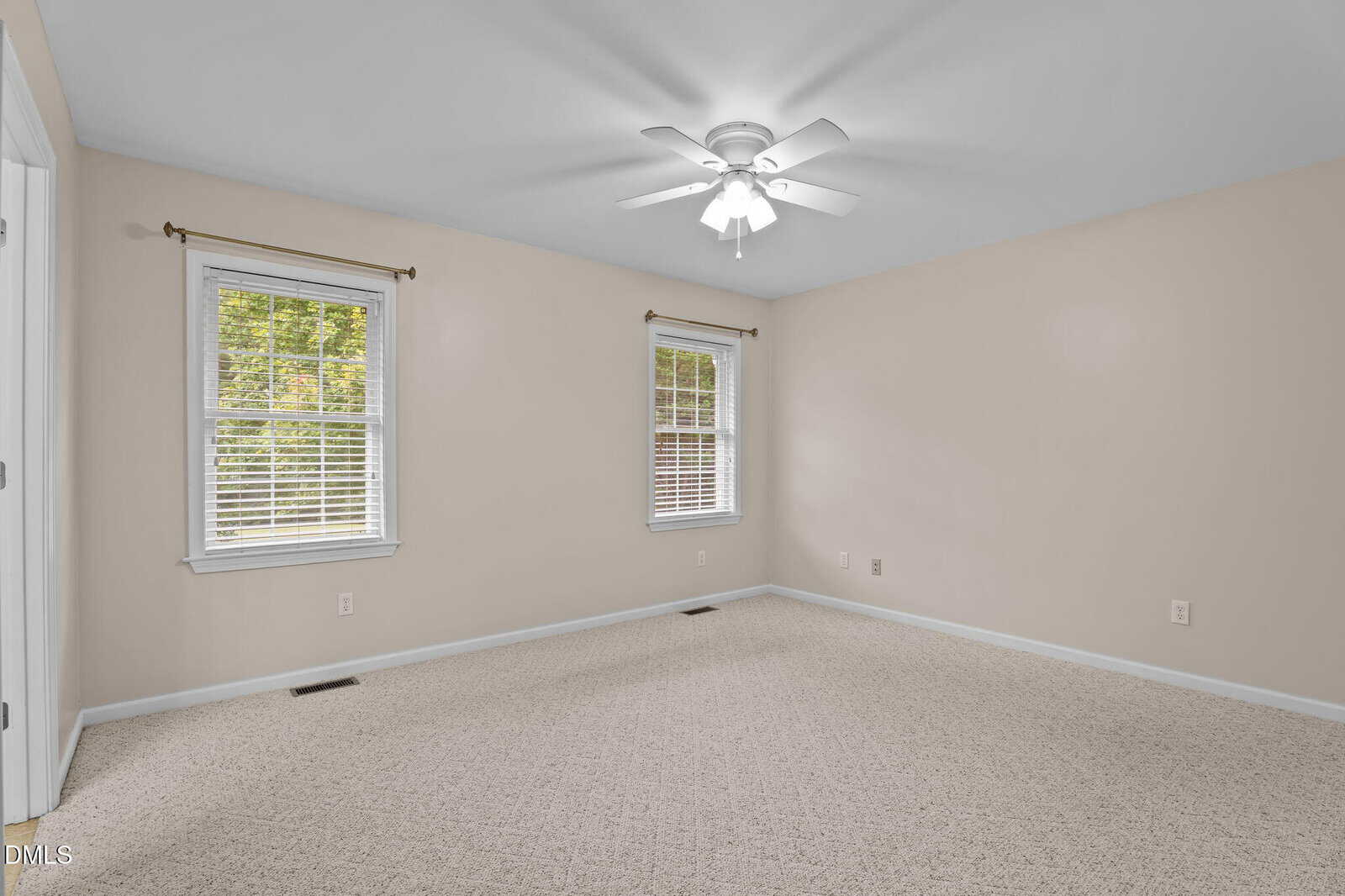 Empty beige-carpeted room with two windows, cream-colored walls, white ceiling fan with lights, and white trim. Daylight comes through the windows, showing green trees outside.