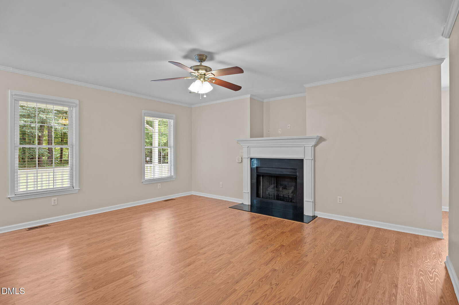 A spacious living room with light beige walls, hardwood floors, two large windows, a ceiling fan with lights, and a white mantel fireplace with a black surround.