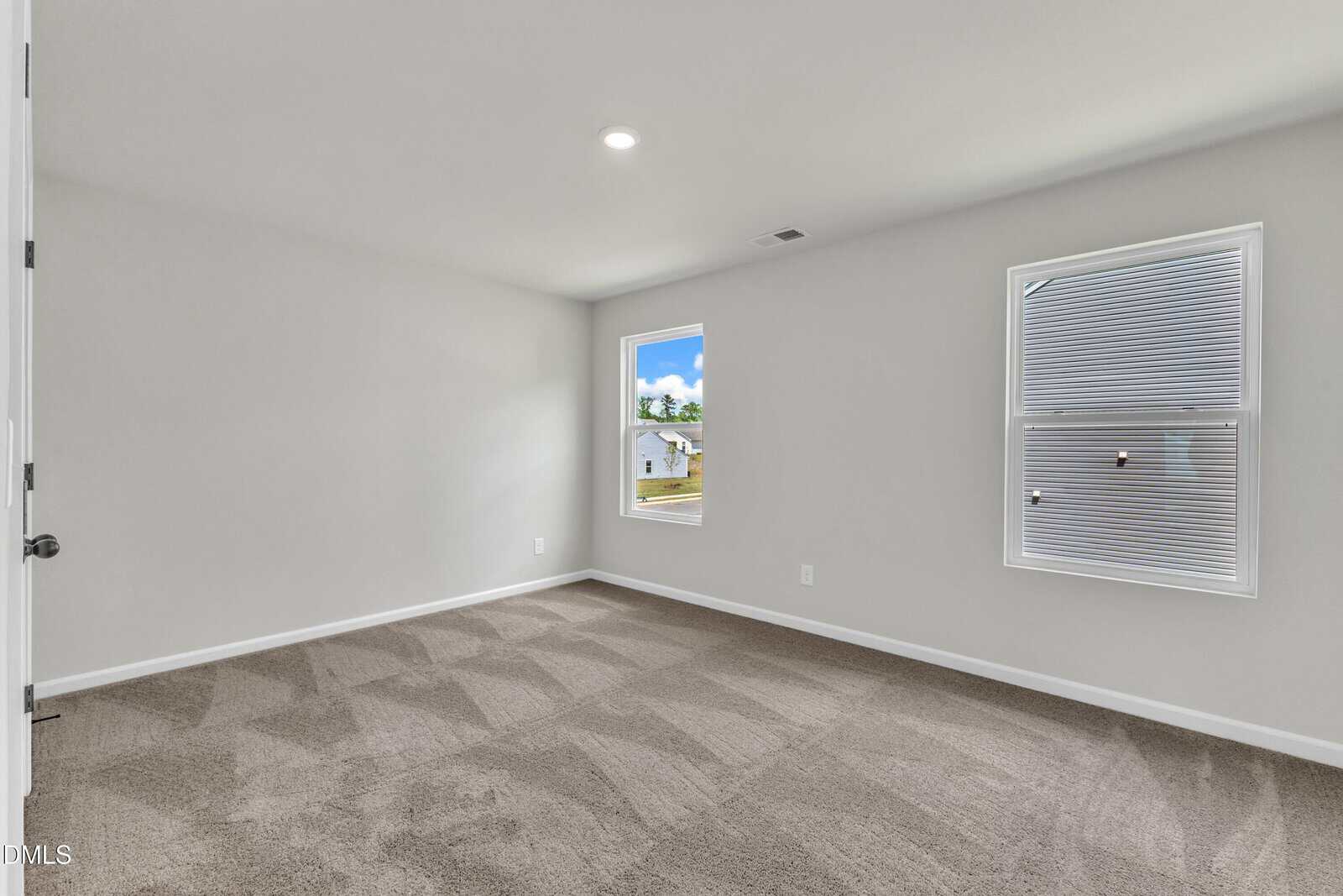 A clean, empty room with light gray walls, beige carpet, two windows with blinds, and a white ceiling with a single recessed light. Natural light comes in through the windows, and part of a house is visible outside.