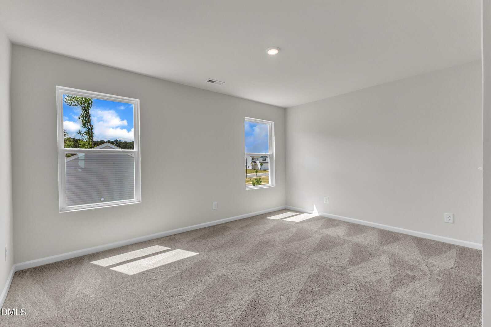Bright, empty room with two large windows, beige carpet, white walls, and a ceiling light. Sunlight streams in, creating patterns on the floor; view of houses and trees outside.
