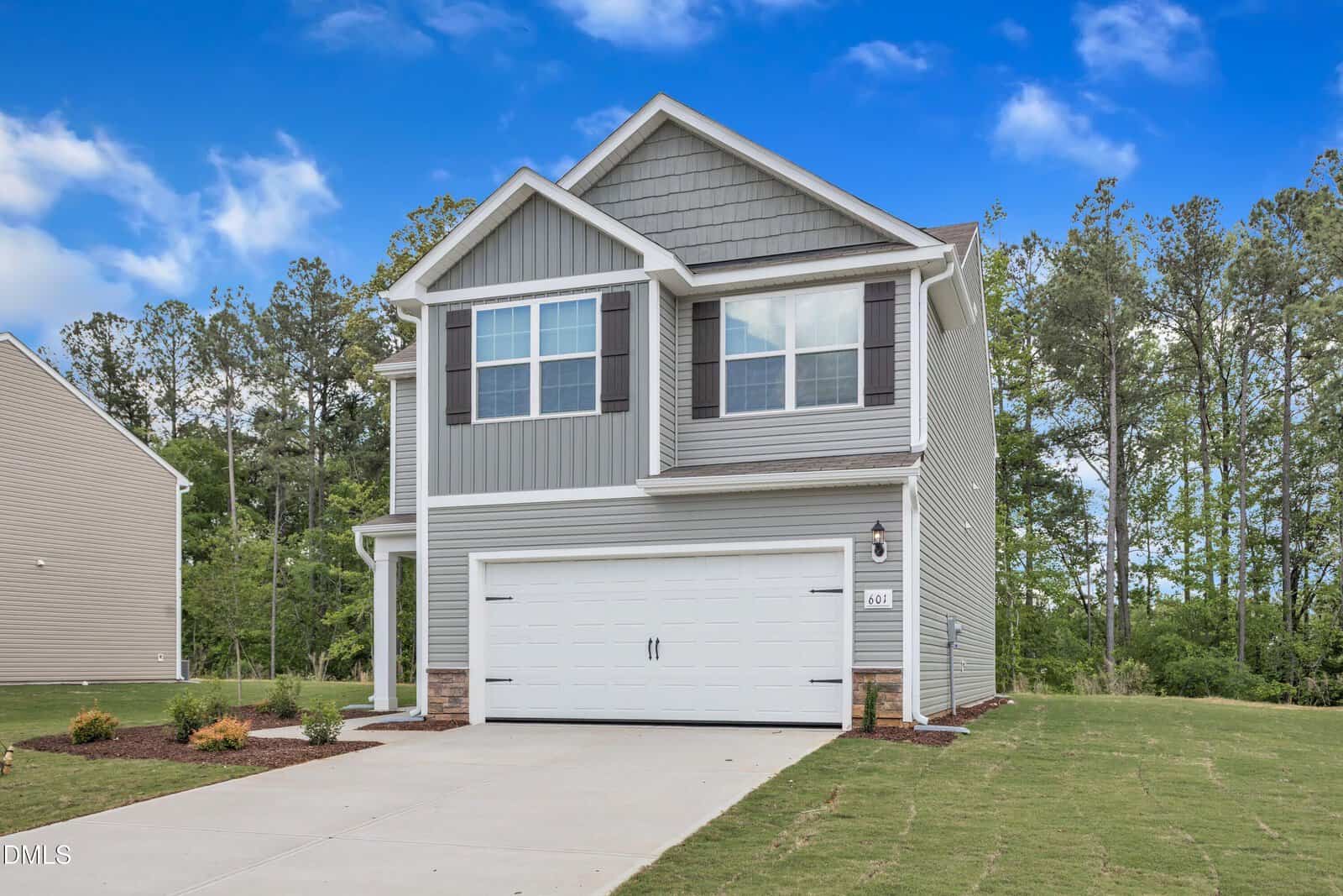 Two-story gray house with white trim, a double garage, and a concrete driveway at 104 Flue Court, Oxford. The newly landscaped yard borders a wooded area and sits under a blue sky with clouds.
