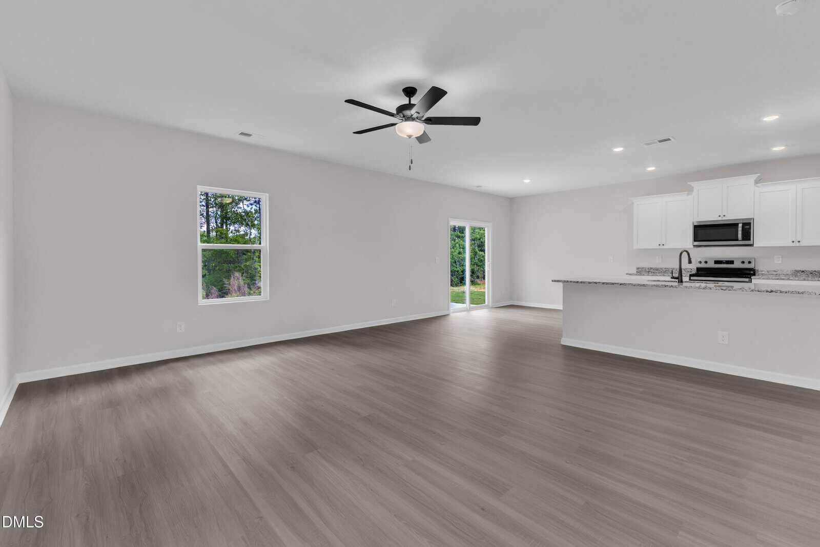 Unfurnished open-concept living space with light gray walls, wood-look flooring, ceiling fan, two windows, a glass door, and a modern kitchen area with white cabinets and stainless steel appliances in the background.