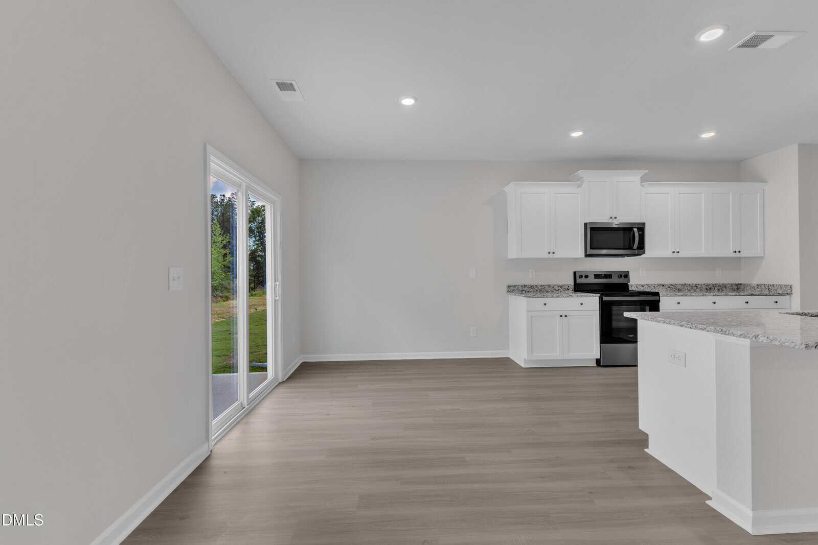 A modern kitchen with white cabinets, granite countertops, stainless steel appliances, and an open dining area. Sliding glass doors open to a grassy outdoor space. The room has light wood floors and white walls.