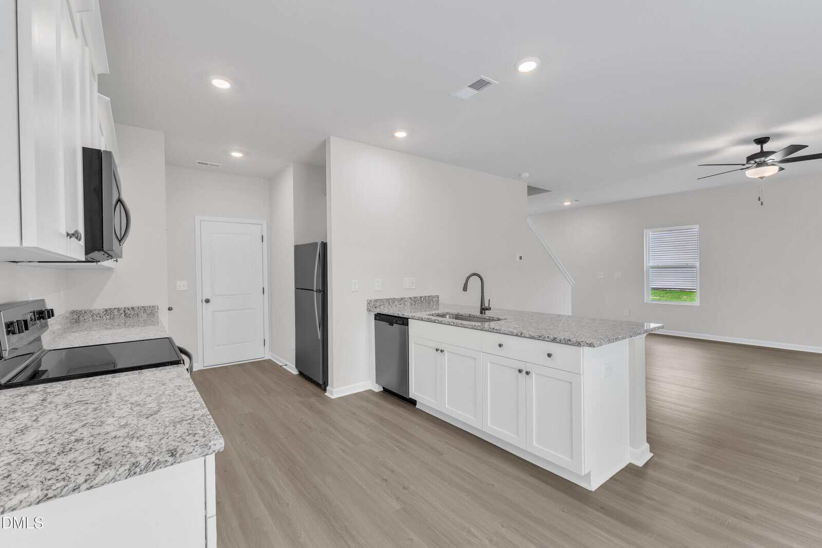Modern kitchen with white cabinets, stainless steel appliances, and granite countertops. Open layout leads to a living area with wood-style flooring, ceiling fan, and a window with blinds.