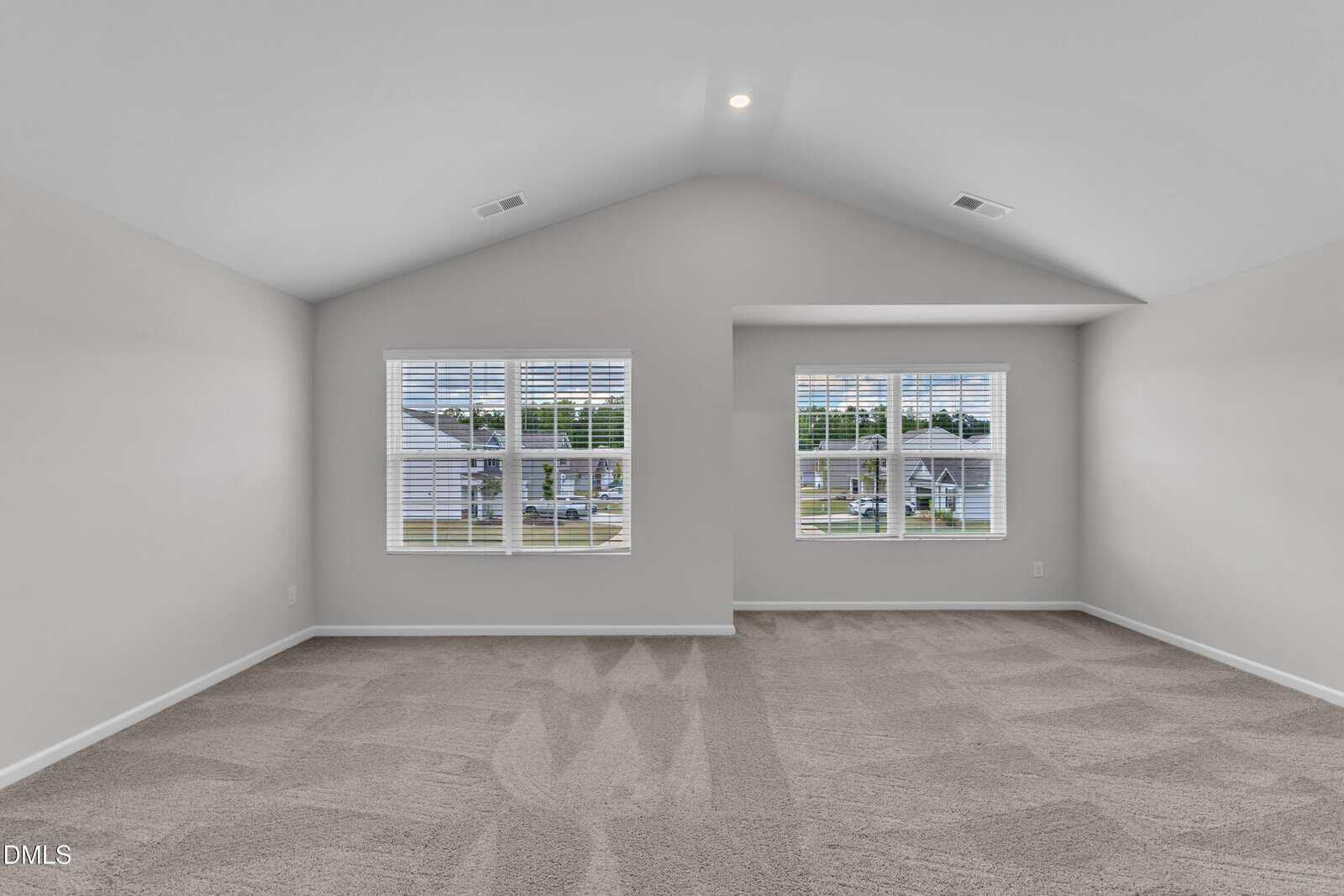 Spacious empty room with light gray walls, vaulted ceiling, and two large windows with white blinds letting in natural light. Beige carpet covers the floor, and suburban houses are visible outside.