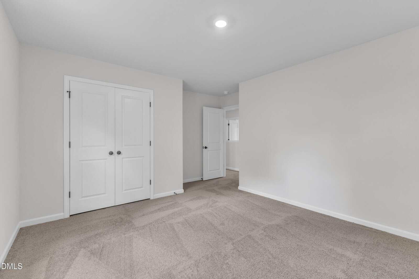 Empty, carpeted room with light beige walls, a ceiling light, a double-door closet, and an open doorway leading to another space. The room is well-lit and has a clean, modern appearance.