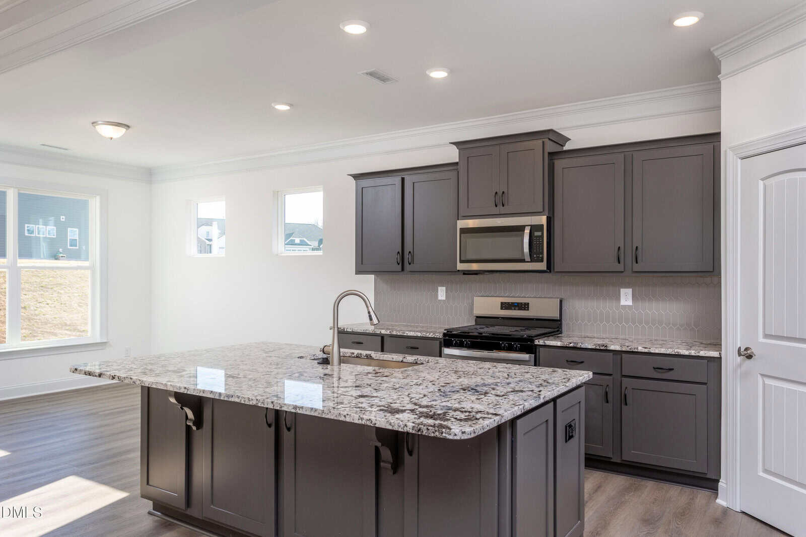 Modern kitchen with dark gray cabinets, stainless steel appliances, a marble-patterned island with a built-in sink, and wood flooring. Large windows let in natural light, creating a bright, open space.