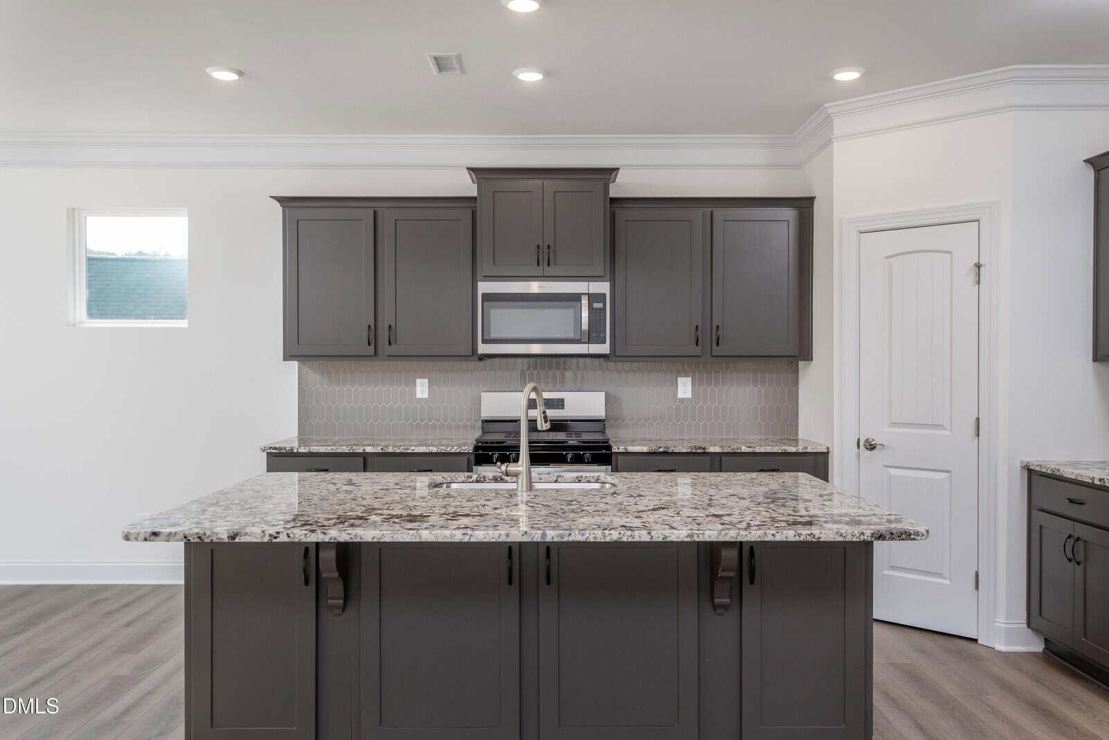 Modern kitchen with gray cabinets, stainless steel appliances, granite countertops, a central island with a sink, and a tile backsplash. There is a window on the left and a white door on the right.