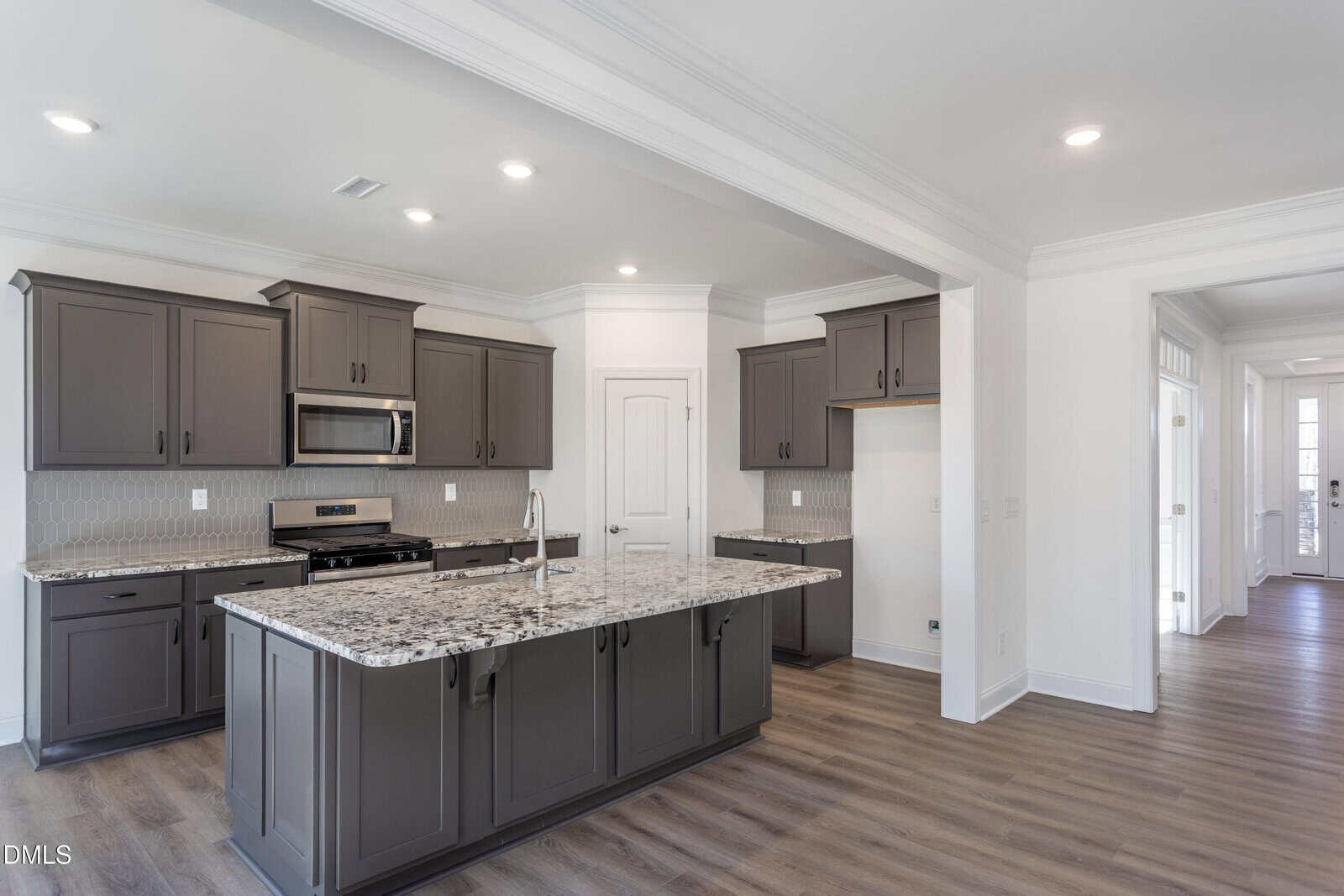 Modern kitchen with dark gray cabinets, stainless steel appliances, a large granite island with a sink, light gray walls, and wood-style flooring. There is ample natural light and an open layout leading to another room.