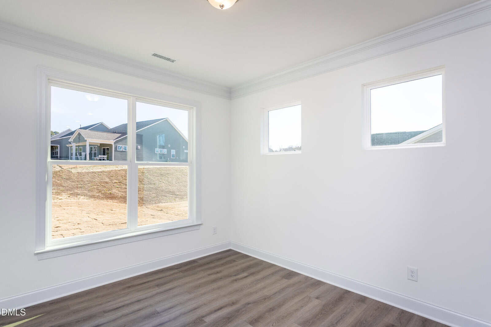 Bright, empty room with white walls, three large windows, and wood-style flooring. Outside, other houses and a dirt yard are visible through the windows. Ceiling light is on.