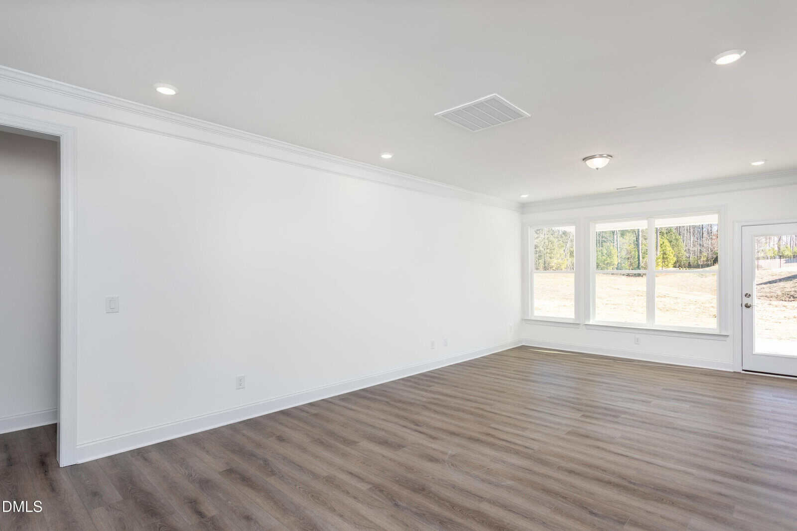 Spacious, empty room with white walls, large windows letting in natural light, recessed ceiling lights, and wood-style flooring. A glass door leads outside to a sunny yard.