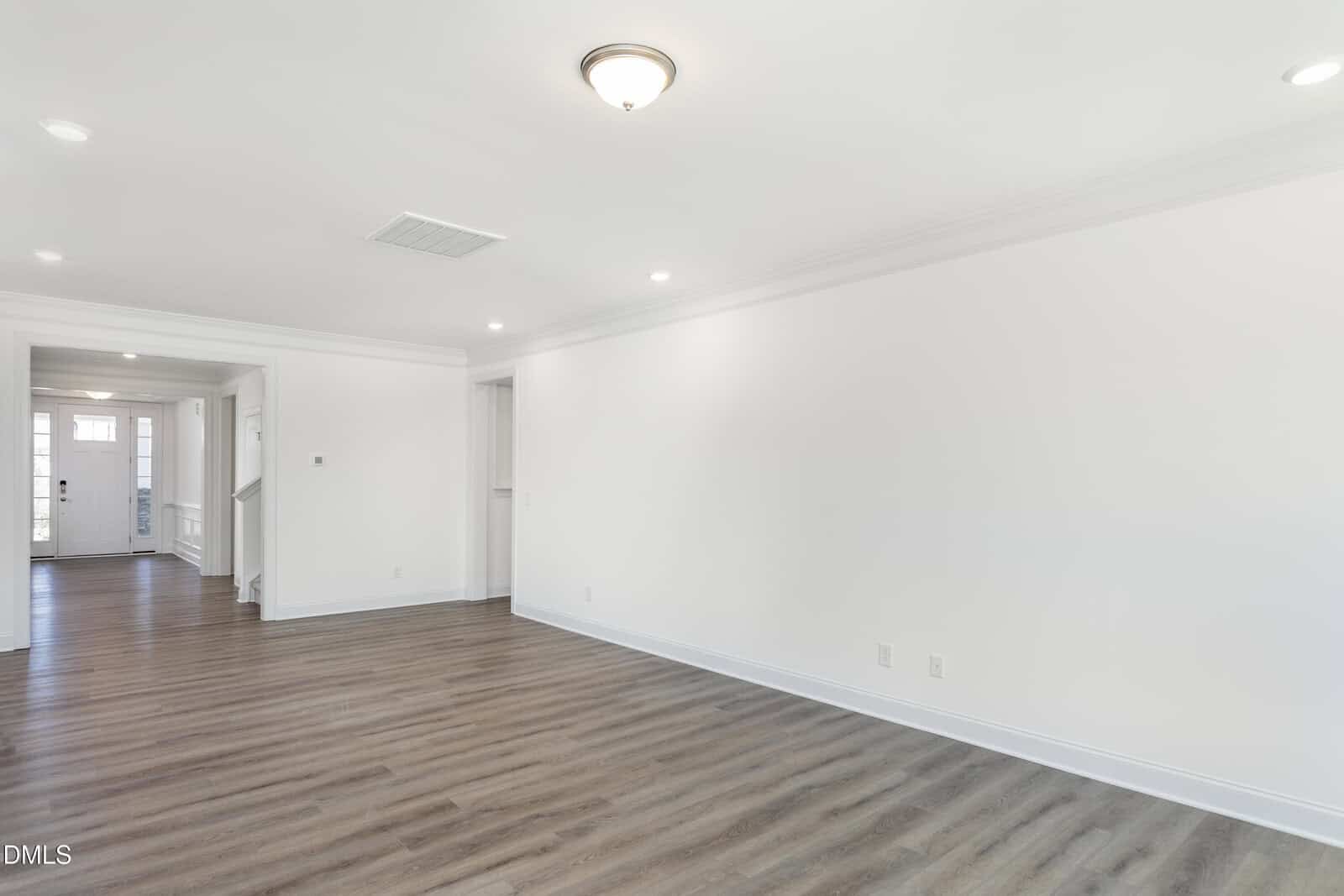 A spacious, empty living room with white walls, recessed lighting, wood-style flooring, and a ceiling light fixture. A hallway leads to the front door in the background.
