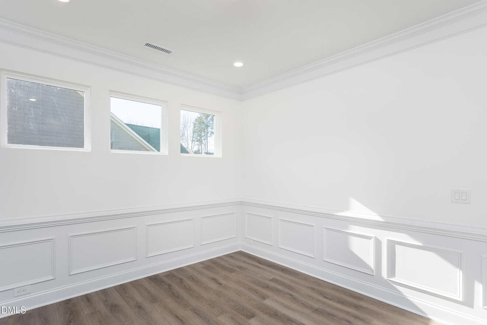 A bright, empty room with white walls, decorative wainscoting, three square windows, and wood-style flooring. Sunlight streams in from the right, illuminating the clean and modern space.