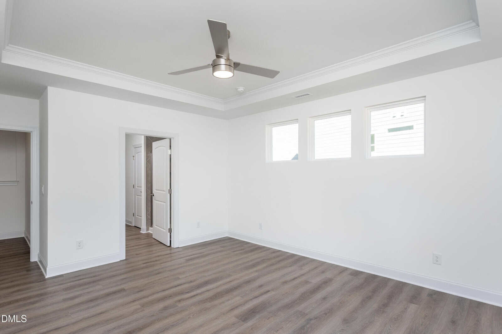 Bright, empty room with light wood floors, white walls, three high rectangular windows, a ceiling fan, and an open doorway leading to another area.