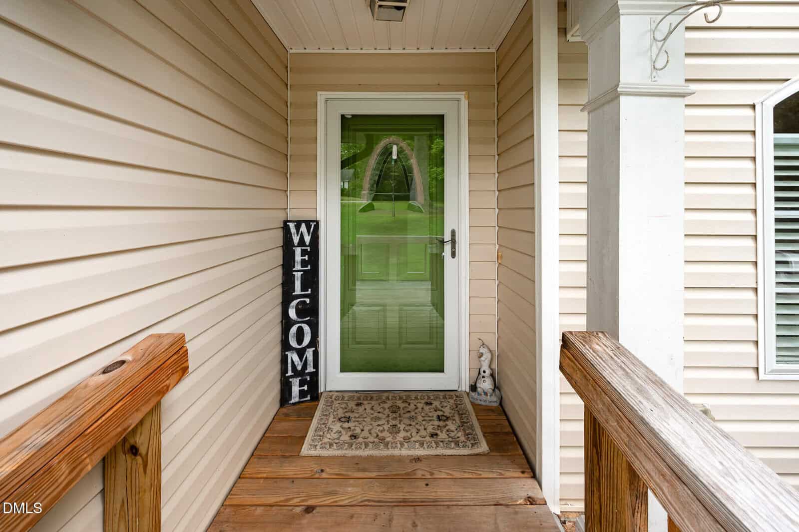 A small front porch with wooden flooring leads to a green door with a glass panel. A doormat and a vertical "WELCOME" sign are by the door, with beige siding on the house walls.