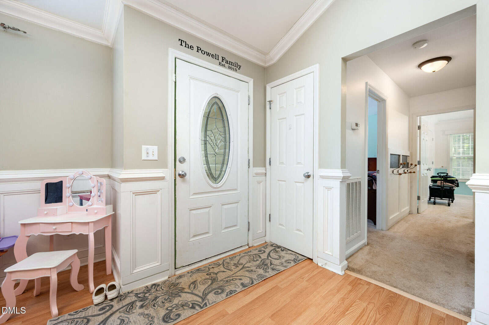 A bright entryway with a glass oval front door, a rug, and white paneled walls. A child's pink vanity set sits to the left. Two doors and a hallway are visible; "The Powell Family" is written above the door.