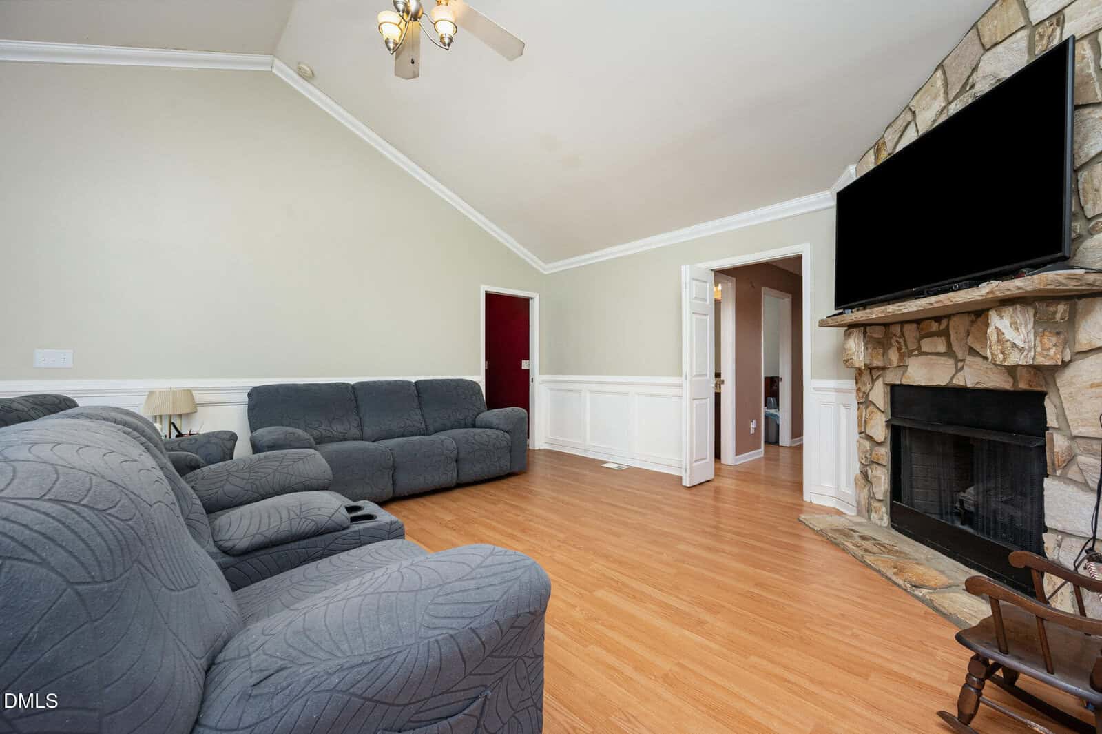 A living room with gray recliner sofas, wood flooring, a stone fireplace with a mounted TV above it, light-colored walls, and a ceiling fan. There is a doorway leading to another room.