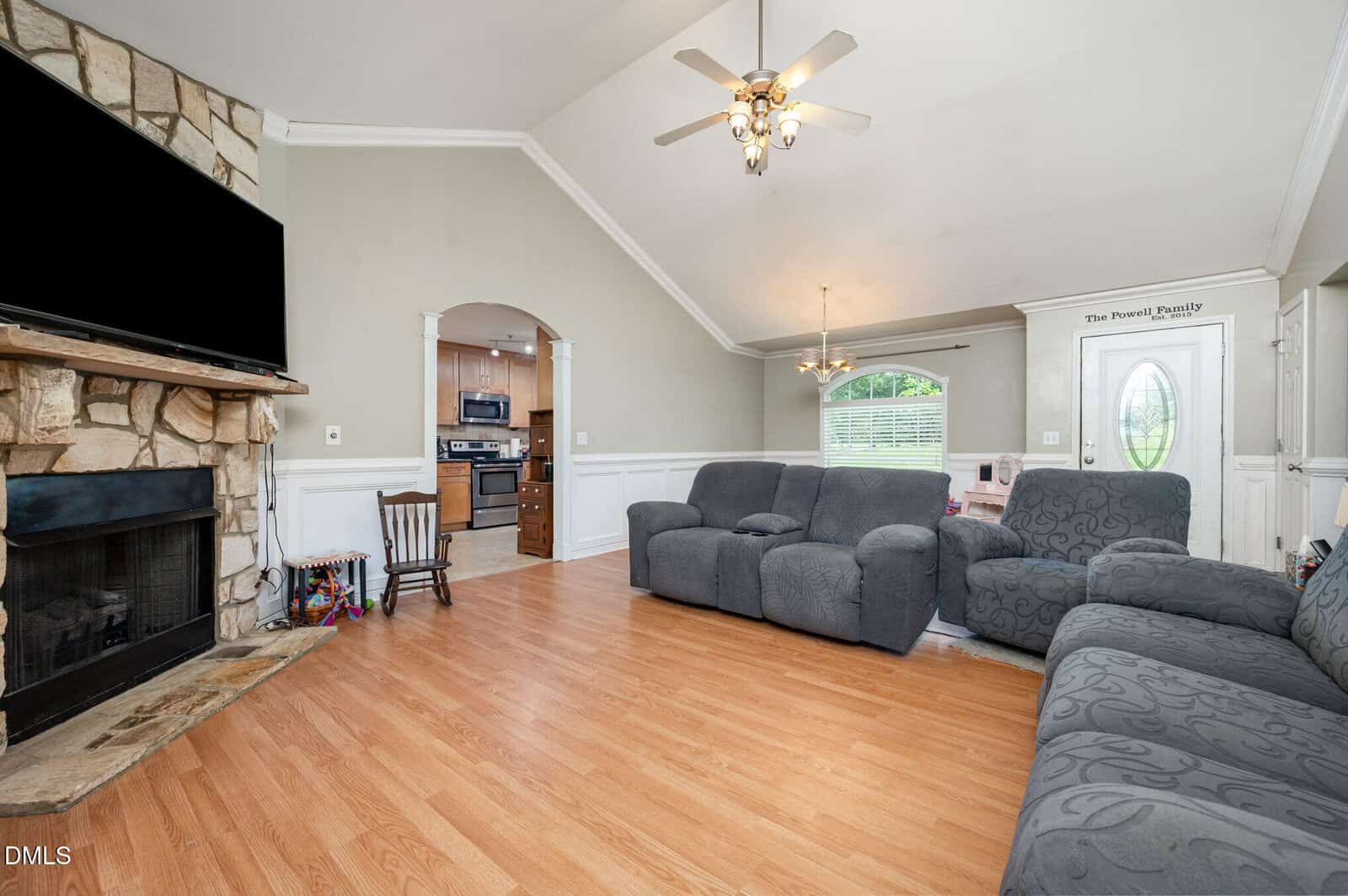 A living room with wood flooring, gray sofas, a stone fireplace with a mounted TV, and a ceiling fan. An arched doorway leads to a kitchen, and a window provides natural light. The front door is visible on the right.