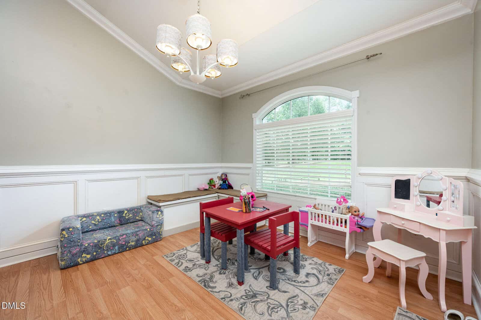 A children's playroom with a small red table and matching chairs on a rug, a floral-patterned sofa, toy vanity set, baby doll crib, and a large window with white blinds letting in natural light.