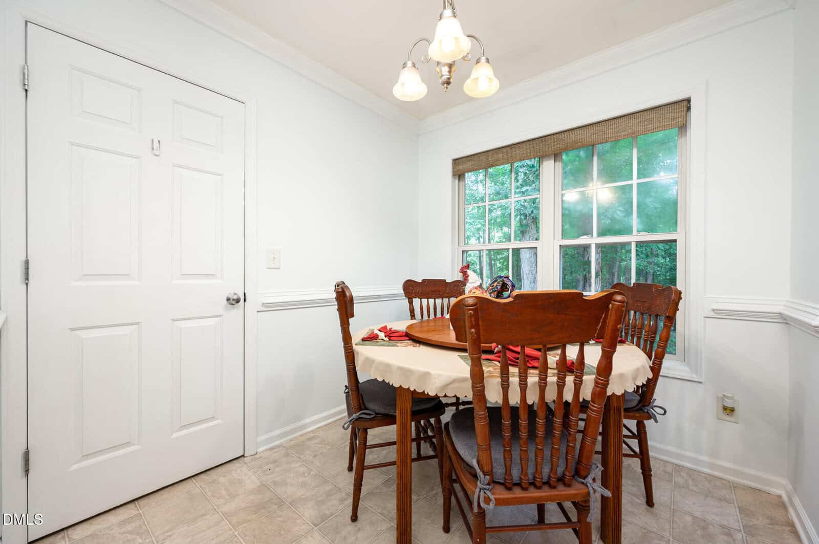 A small dining area features a round wooden table with a beige tablecloth, surrounded by four wooden chairs, positioned near a window with a view of greenery and next to a closed white door.