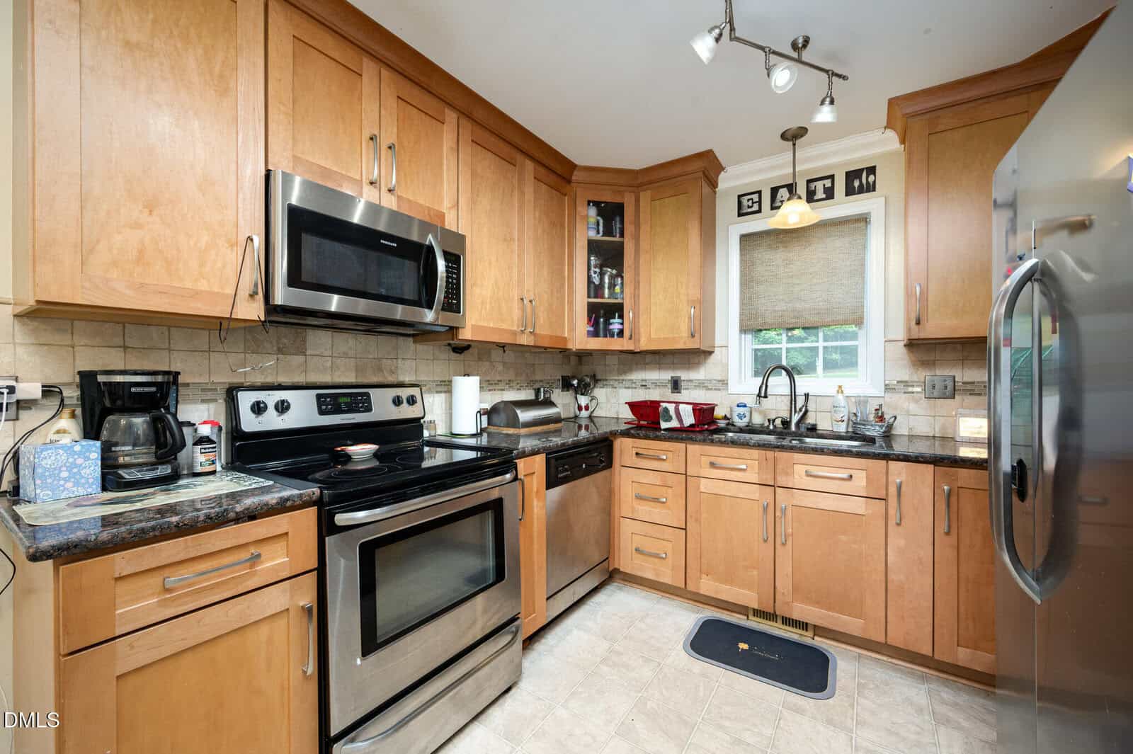 A kitchen with light wood cabinets, stainless steel appliances, a microwave above an electric stove, a refrigerator, a coffee maker, and a window above the sink. The counters have various small kitchen items.
