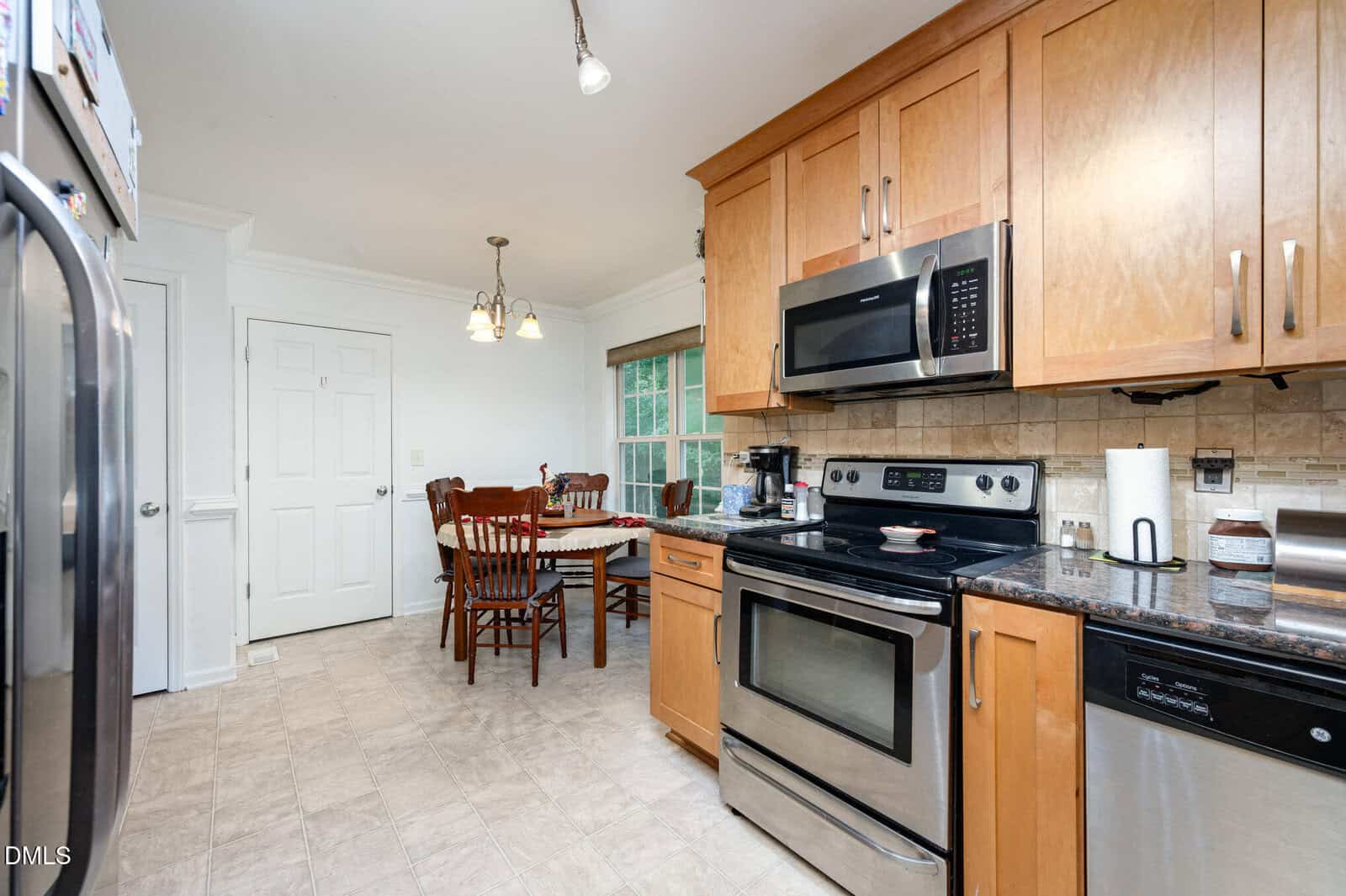 Modern kitchen with stainless steel appliances and wood cabinets, adjacent to a dining area with a wooden table and chairs by large windows, under a hanging light fixture.