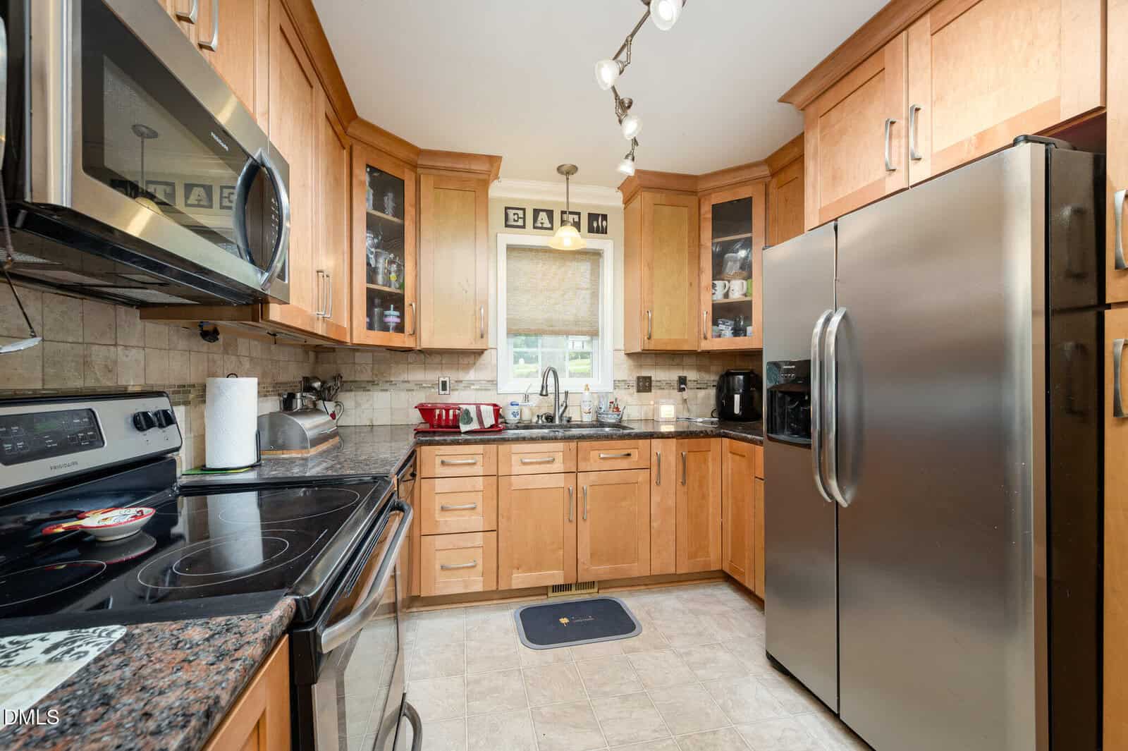 Modern kitchen with wood cabinets, stainless steel appliances, black granite countertops, and tiled backsplash. Various small appliances and utensils are on the counters. A window with a shade is above the sink.