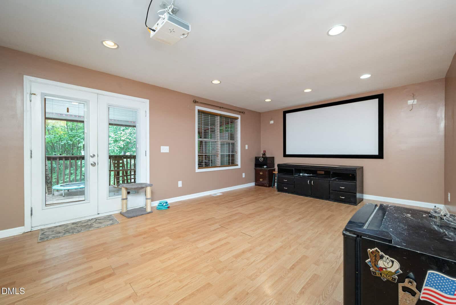Spacious room with light wood floors, beige walls, a large projector screen, media console, window, double glass doors to a deck, pet bowls, and scattered items on a black piece of furniture.