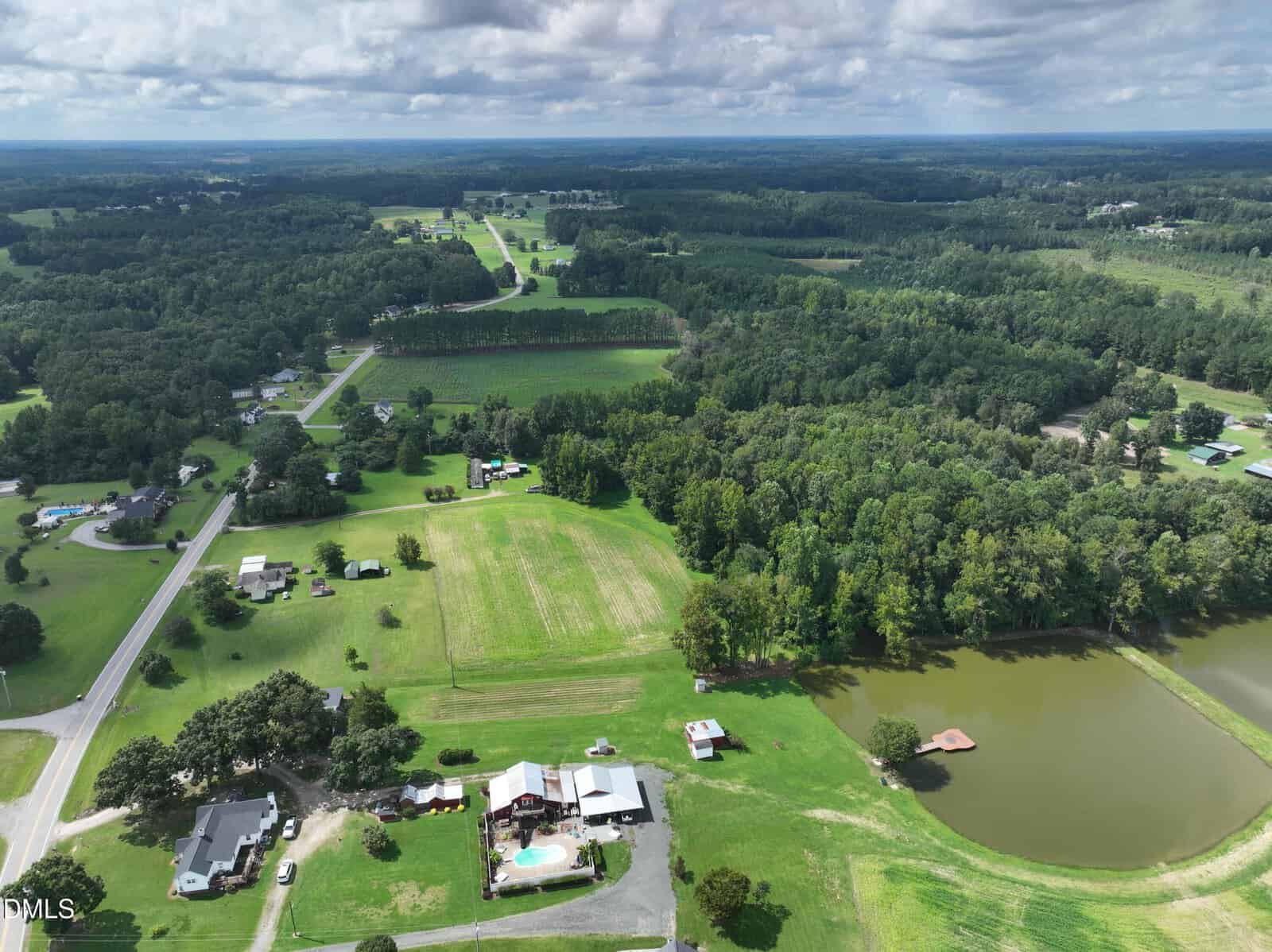 Aerial view of a rural landscape with green fields, scattered houses, a small lake with a dock, and a swimming pool near one home; bordered by forests and intersected by roads under a cloudy sky.