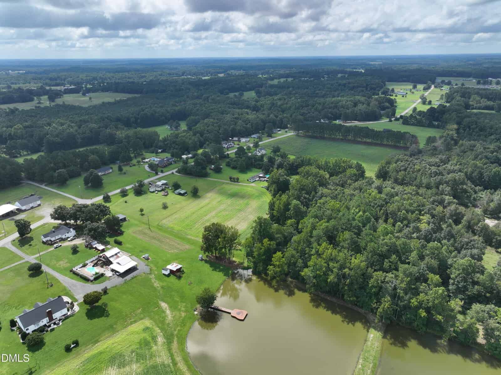 A rural landscape with scattered houses, open green fields, dense trees, and a pond with a small dock visible. Roads connect properties, and the sky is partly cloudy. The horizon is filled with forest and open land.