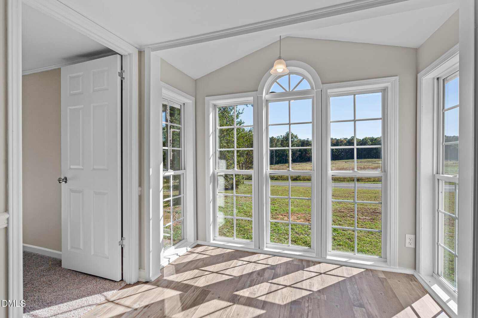 A bright sunroom with large, white-trimmed windows and a glass door, allowing natural light to fill the space. The room features light wood flooring and a view of a grassy field and trees outside.