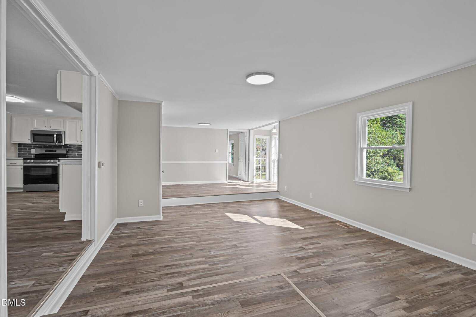 A spacious, empty room with wood flooring, white walls, and a window on the right. The room opens to a kitchen with white cabinets and stainless steel appliances, and a dining area with glass doors leading outside.