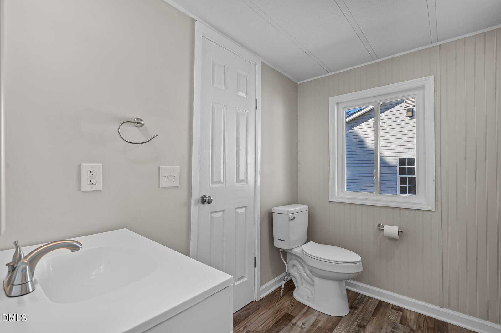 A bright, clean bathroom with a white sink, toilet, towel ring, and a window showing the exterior of a house. The walls are light beige with wood paneling, and the floor has a wood-like finish.