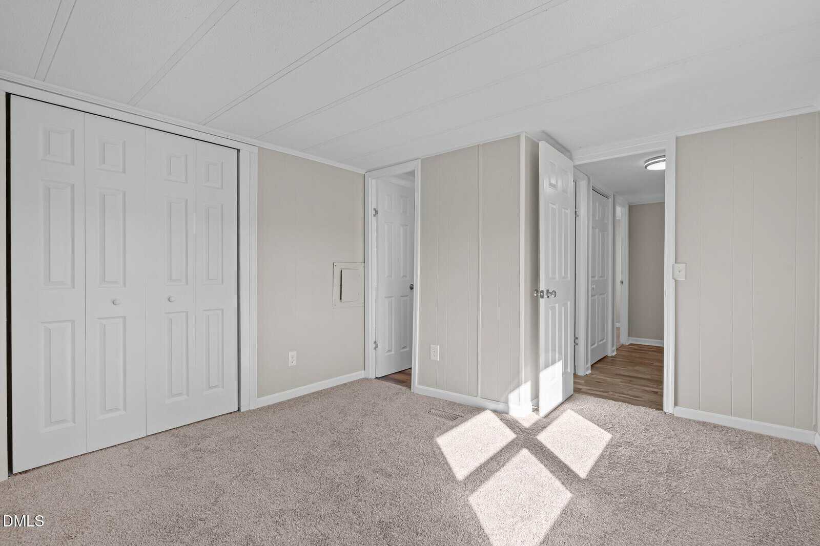 Bright, empty room with beige carpet, light-colored walls, multiple white doors, and a closet. Sunlight creates a large X-shaped shadow on the floor.