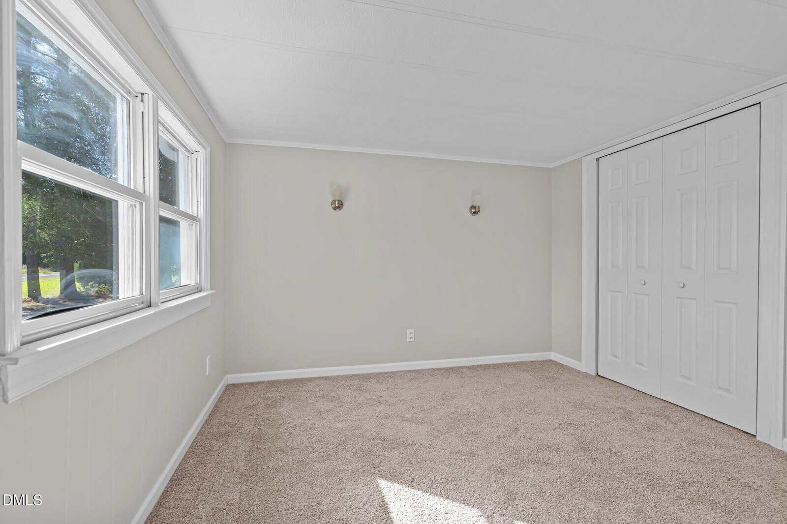 Bright, empty room with beige carpet, light gray walls, two windows on the left, two wall sconces, and a white double-door closet. Sunlight streams in through the windows.