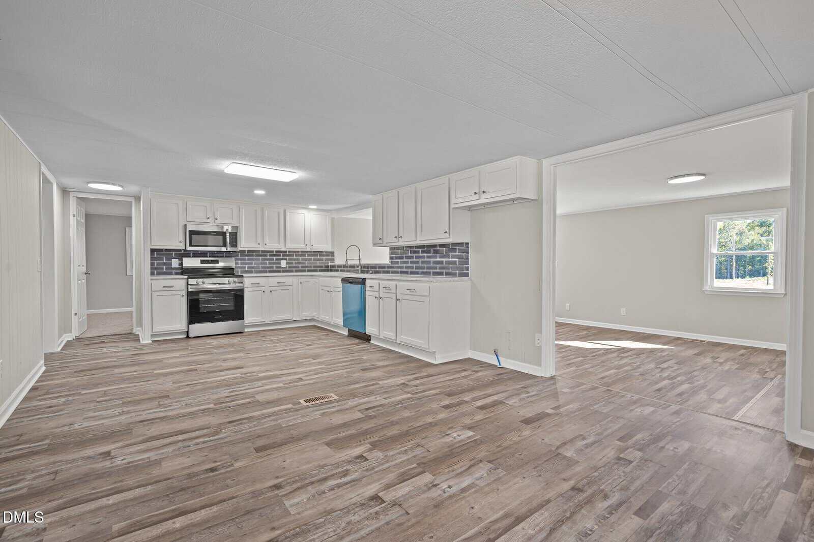 A spacious, open-concept kitchen and living area with wood-style flooring, white cabinets, stainless steel appliances, a blue dishwasher, and a window letting in natural light. A second room is visible through a wide doorway.