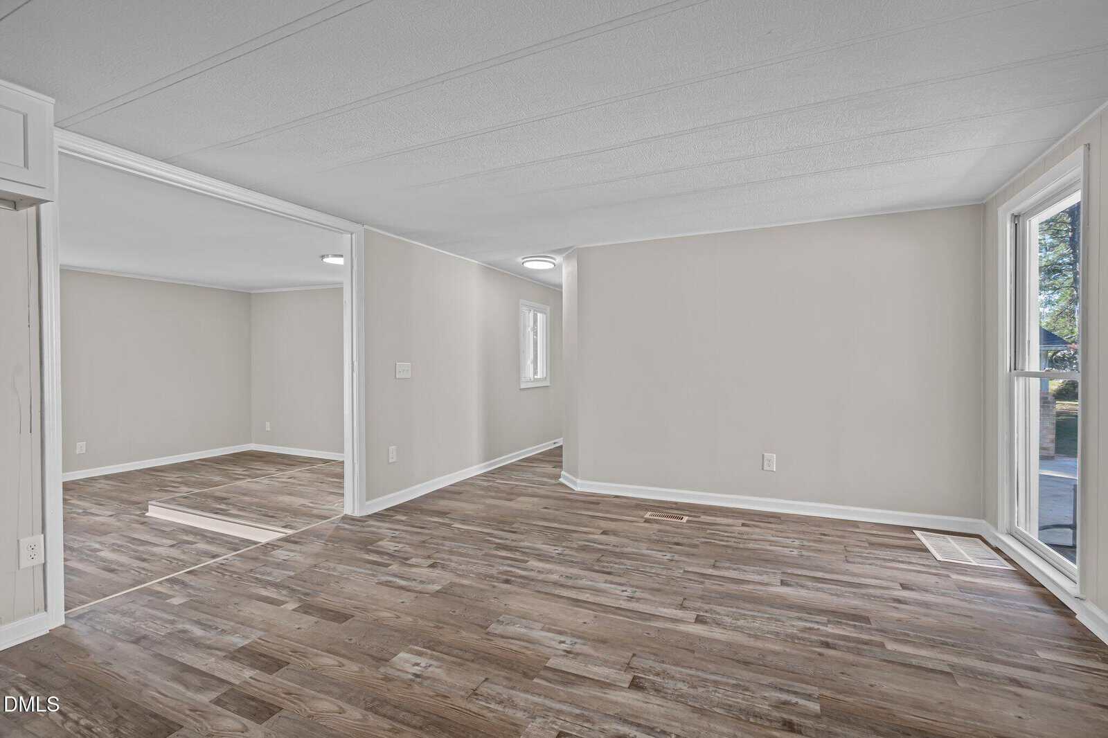Empty room with light beige walls, white trim, and wood-look vinyl flooring. Large windows let in natural light. The space is open with visible doorways connecting multiple rooms.