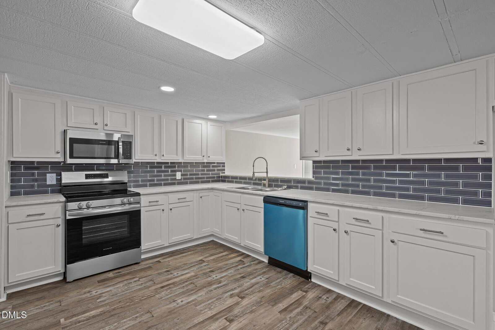 Modern kitchen with white cabinets, gray subway tile backsplash, stainless steel appliances, electric stove, built-in microwave, and a dishwasher with a blue protective film. Wood-style flooring is visible throughout.