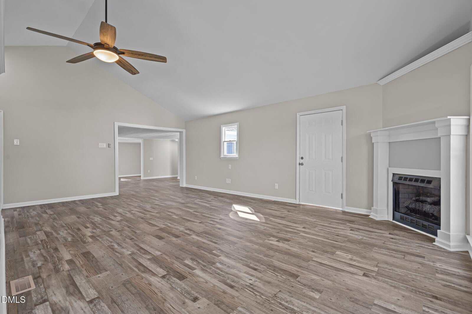 Spacious empty living room with light-colored walls, vaulted ceiling, wood-look flooring, a ceiling fan, a small window, white doors, and a fireplace with a decorative mantel in the corner.